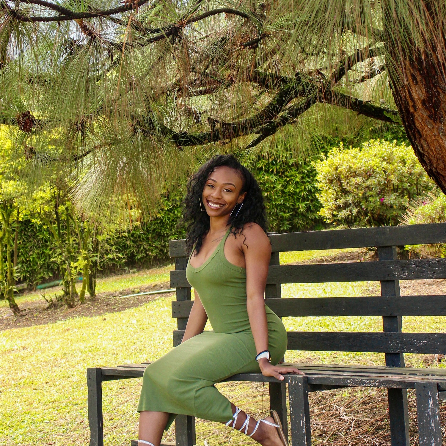 A woman wearing a green sleeveless dress sits on a dark wooden park bench under a pine tree, with grass and shrubs in the background.