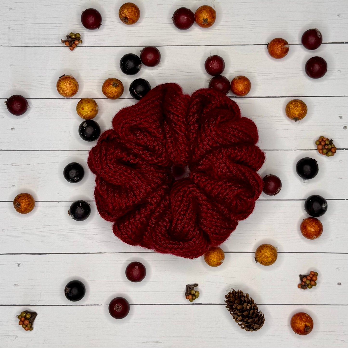 Knitted maroon scrunchie surrounded by small fruits and pine cones on a white wooden background