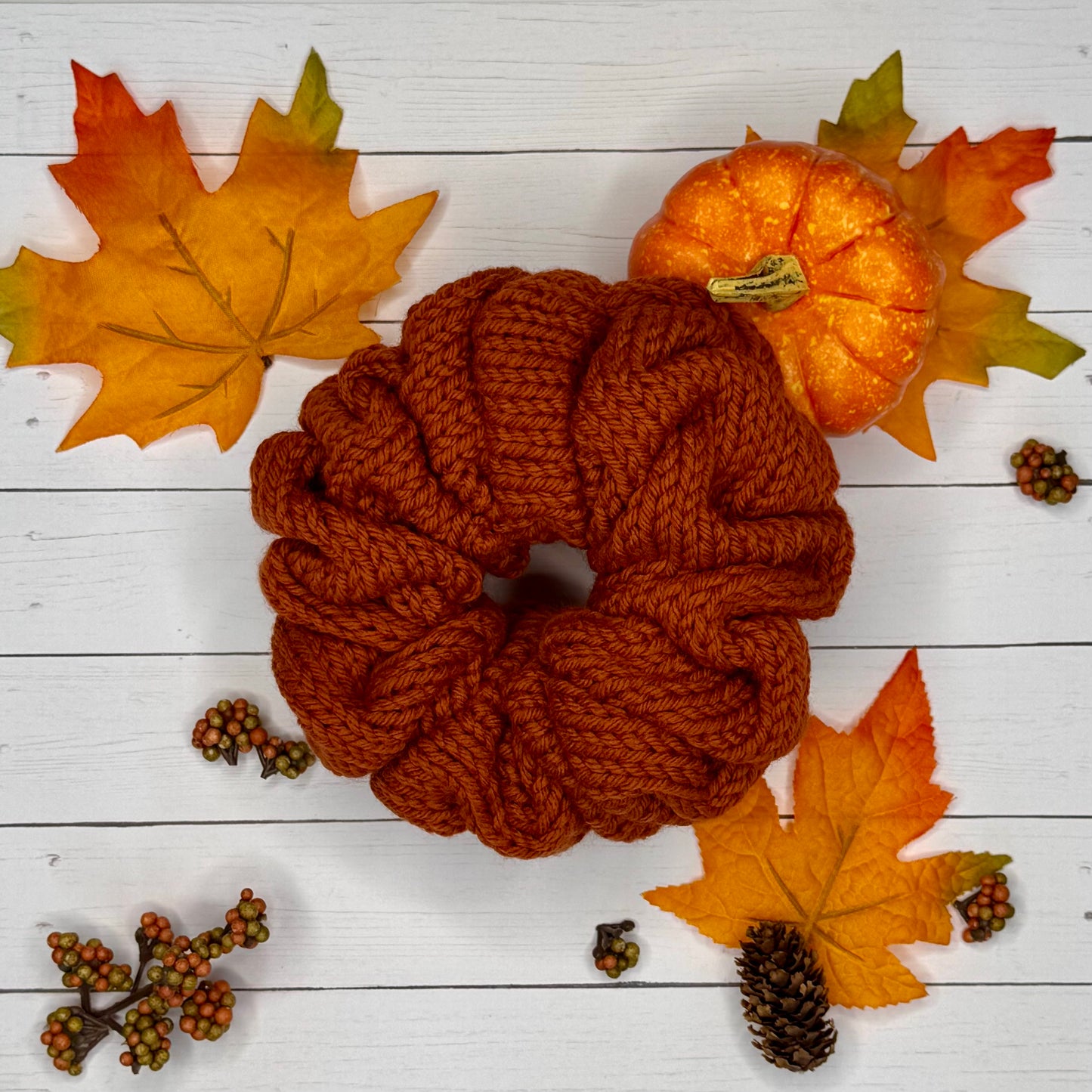 Brown knitted scrunchie with autumn leaves and a small pumpkin on a white wooden background