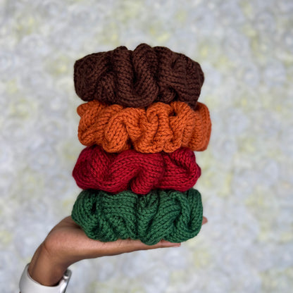 Stack of hand knit jumbo scrunchies in brown, orange, red, and green held against a textured background.