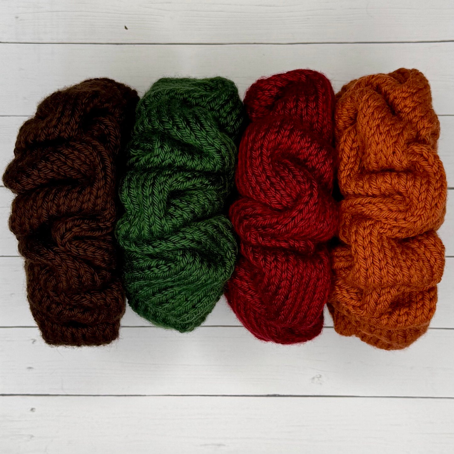 Four scrunchies in brown, green, red, and orange on a white wooden surface