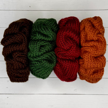 Four scrunchies in brown, green, red, and orange on a white wooden surface